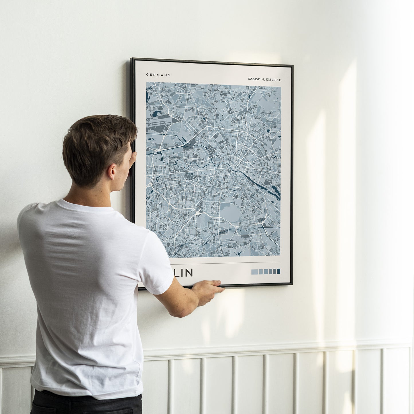 A man in a white t-shirt is shown from behind, holding a framed poster of a blue-gray map of Berlin, Germany, against a white wall with wainscoting.
