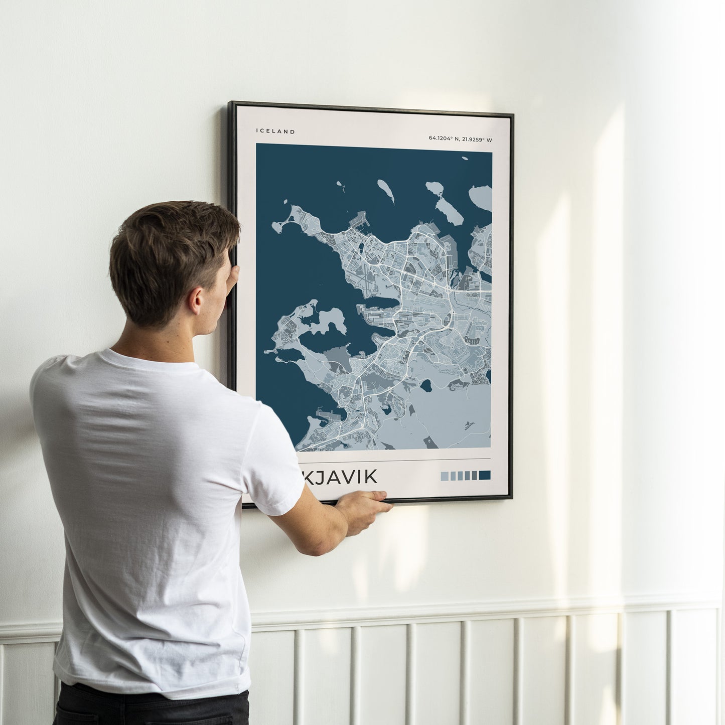 A man in a white t-shirt is shown from behind, holding a framed map print of Iceland's capital, Reykjavík, against a white wall.