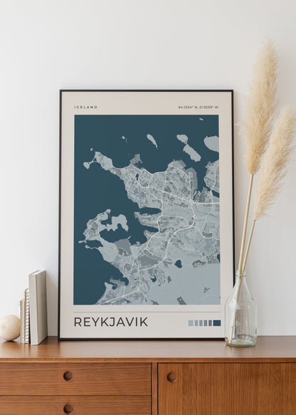 A framed minimalist map print of Reykjavík, Iceland, displayed on a wooden cabinet against a white wall, styled with books, a vase of pampas grass, and a small decorative object.