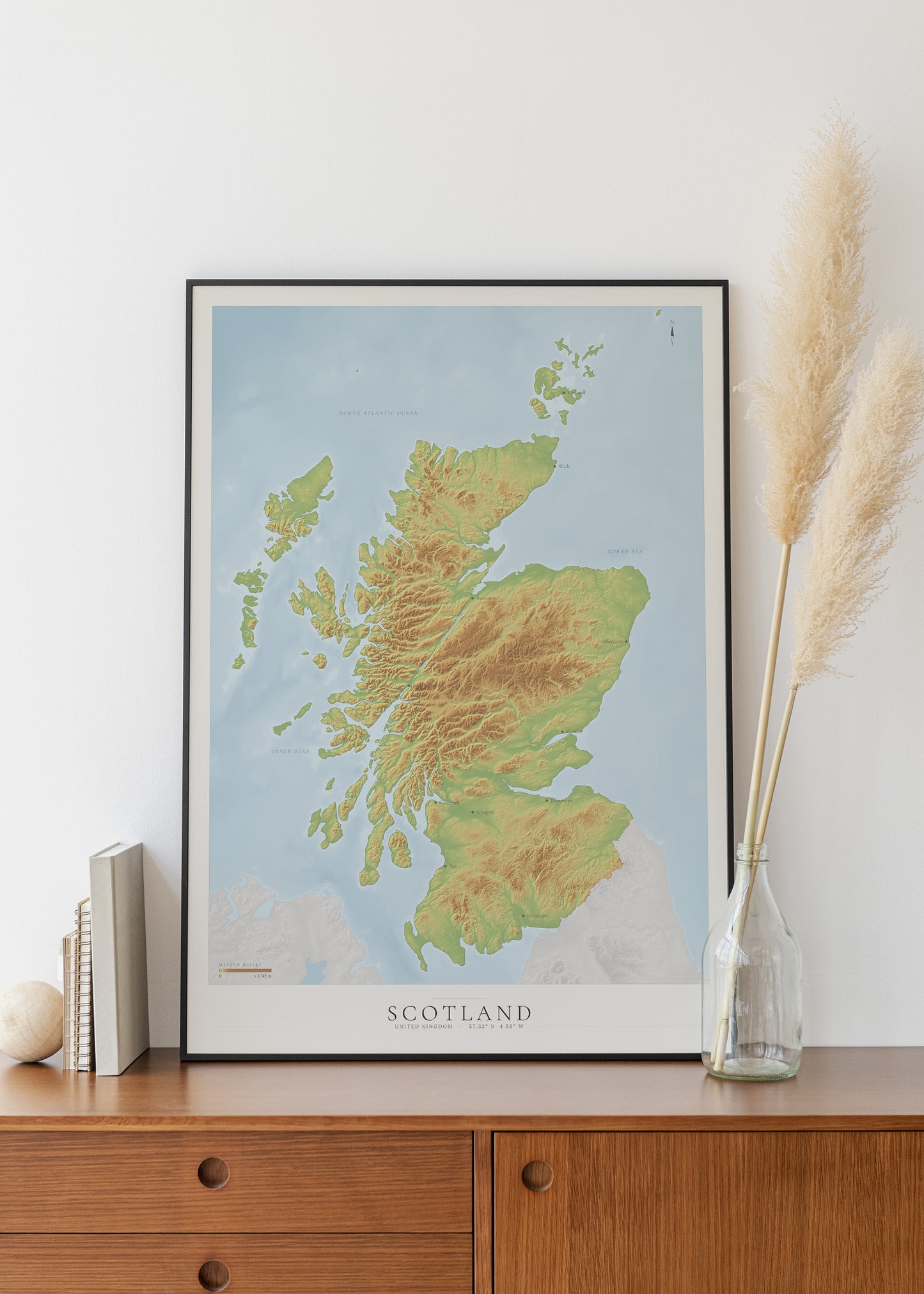 A framed topographical map print of Scotland, displayed on a wooden cabinet against a white wall, styled with books, a small vase, and dried pampas grass.