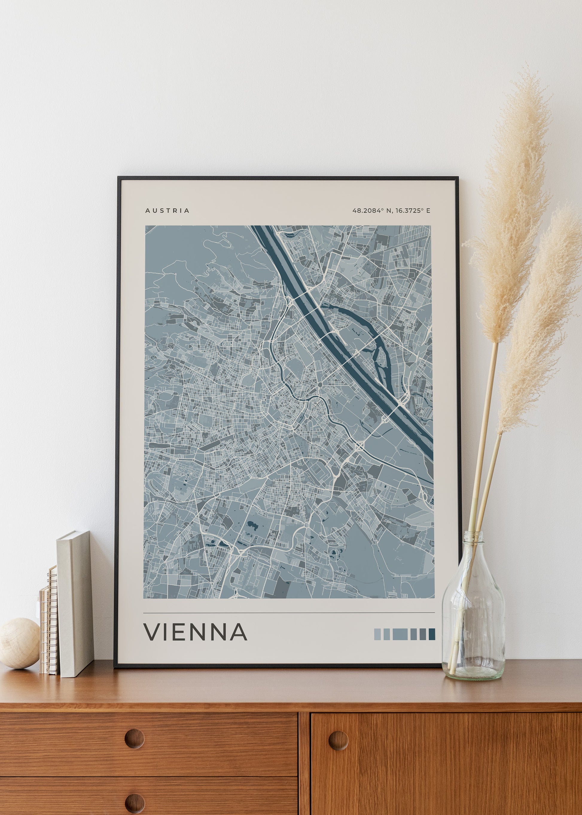 A framed minimalist map print of Vienna, Austria, styled on a wooden sideboard next to a glass vase with pampas grass.
