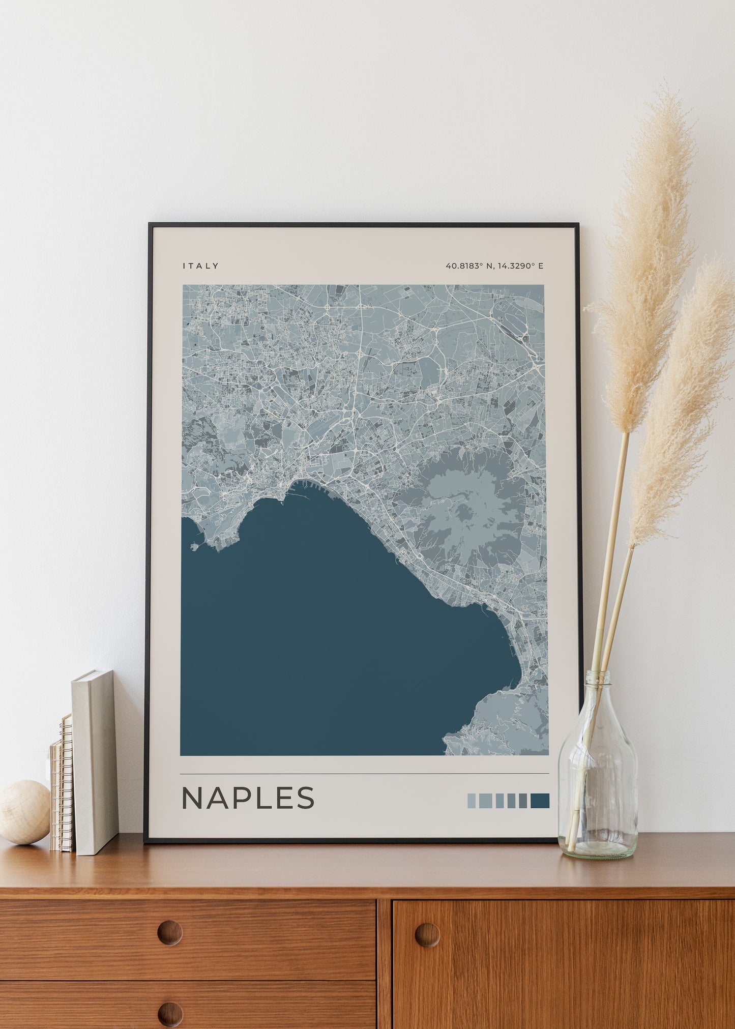 A framed Naples city map print with a dark blue sea area and light blue land, displayed on a wooden cabinet against a white wall with books and a vase of pampas grass.