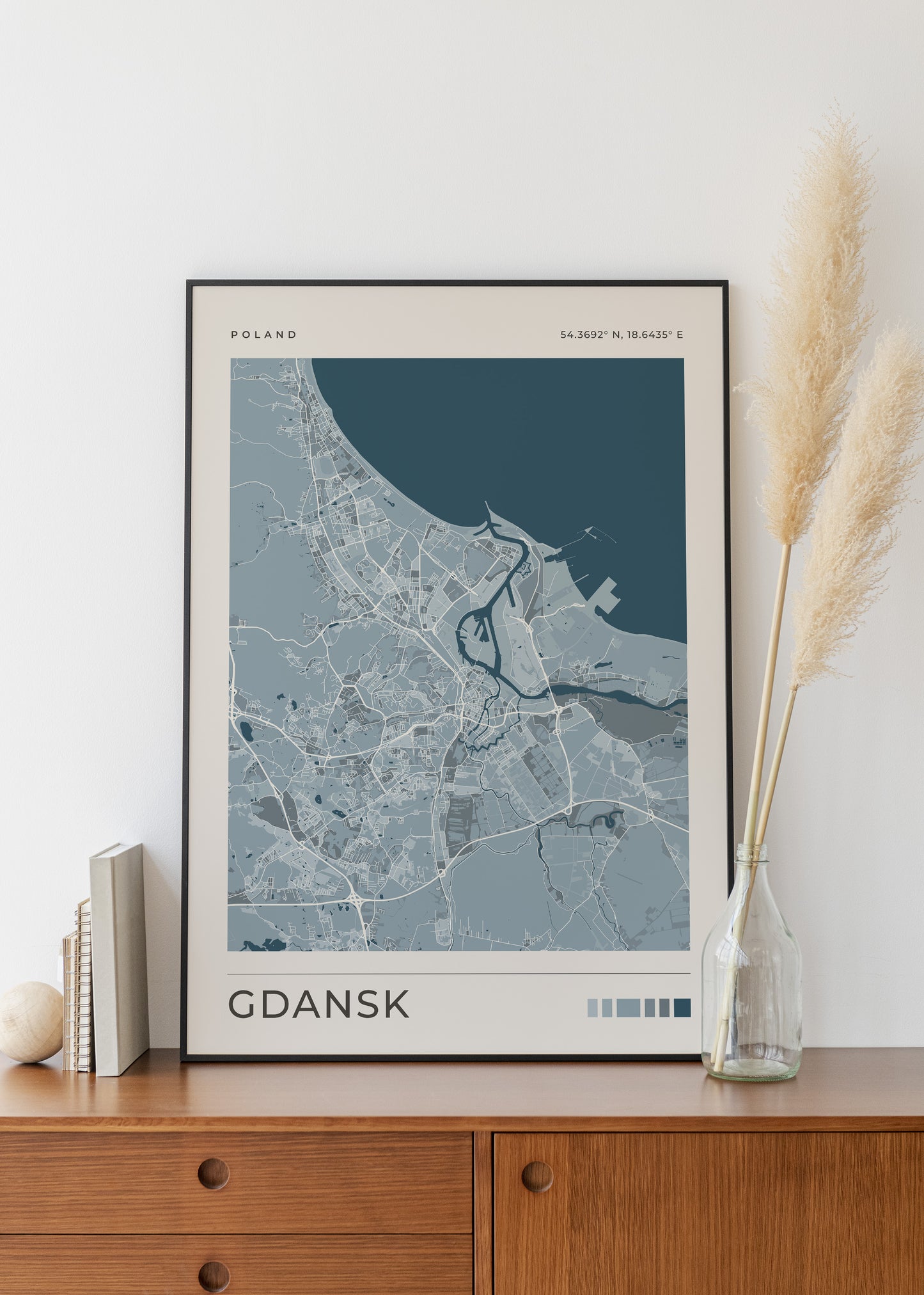 A framed minimalist map print of Gdańsk, Poland, styled on a wooden sideboard next to a glass vase with pampas grass and a small book stack.