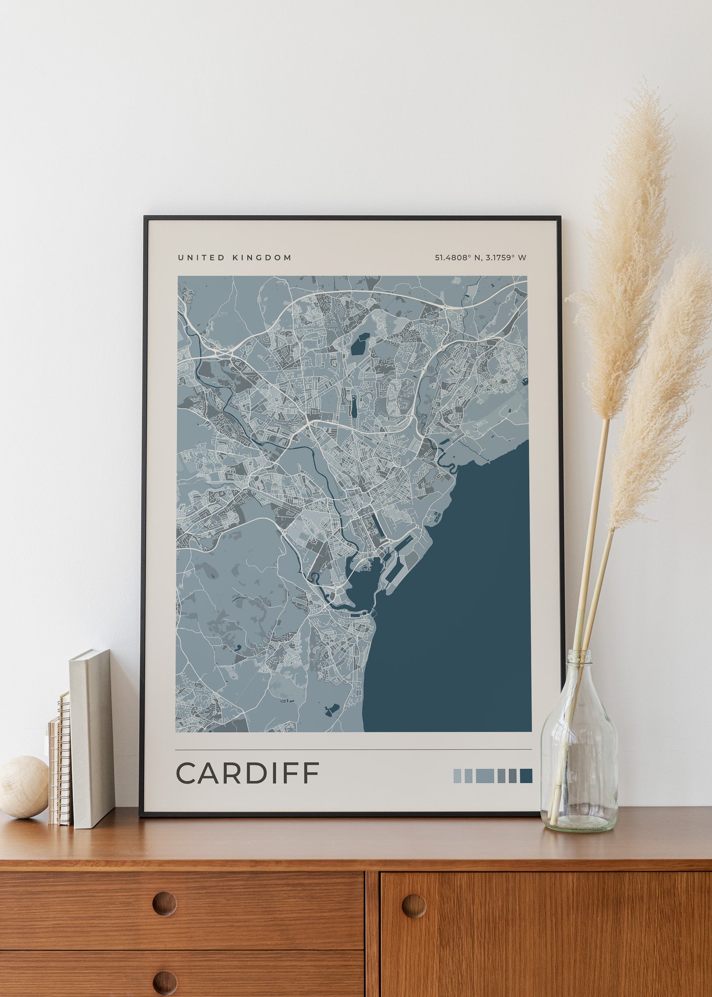 A framed minimalist map print of Cardiff, United Kingdom, styled on a wooden sideboard next to a glass vase with pampas grass.
