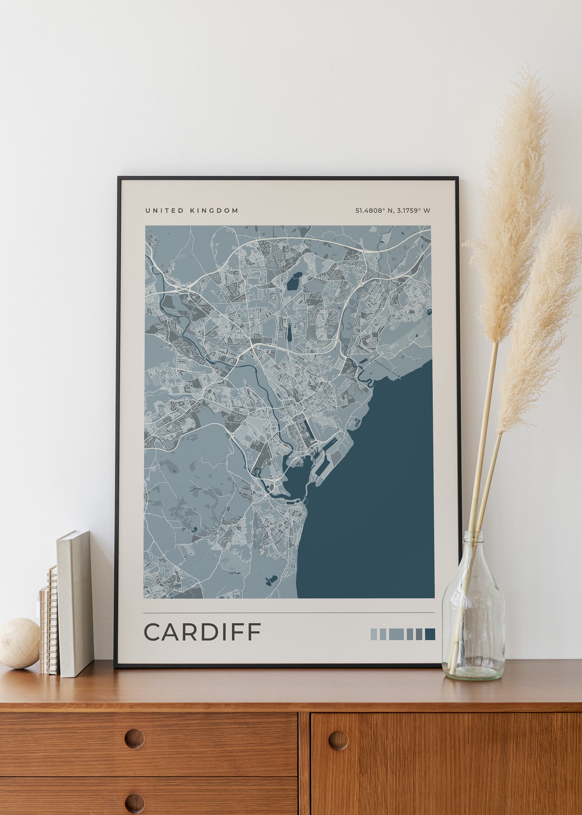 A framed minimalist map print of Cardiff, United Kingdom, styled on a wooden sideboard next to a glass vase with pampas grass.