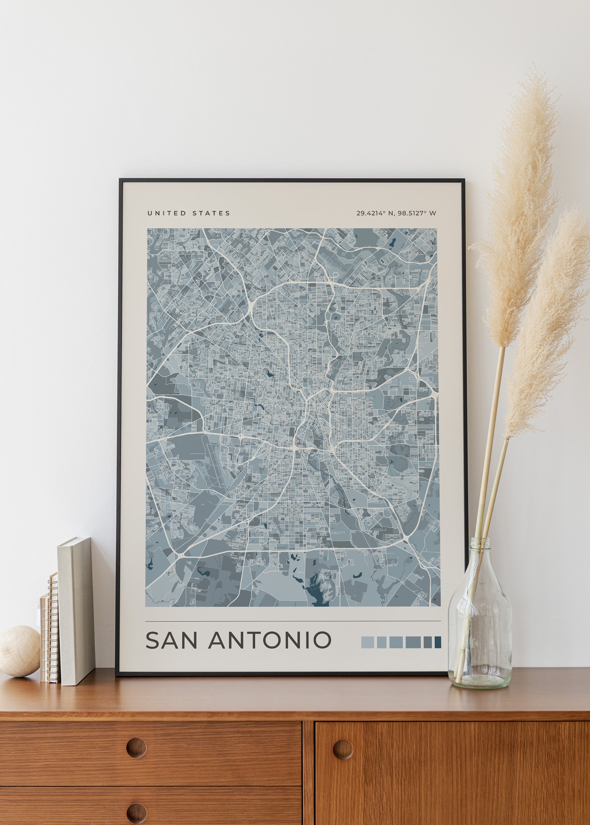 A framed San Antonio city map print with a black frame, leaning on a wooden cabinet against a white wall, with books on the left and a glass vase with dried pampas grass on the right.