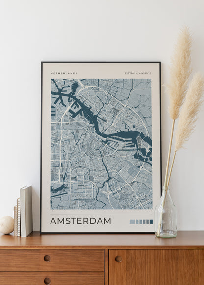 A framed Amsterdam city map poster with a black frame, leaning on a wooden sideboard against a white wall, styled with books, a small vase, and dried pampas grass.