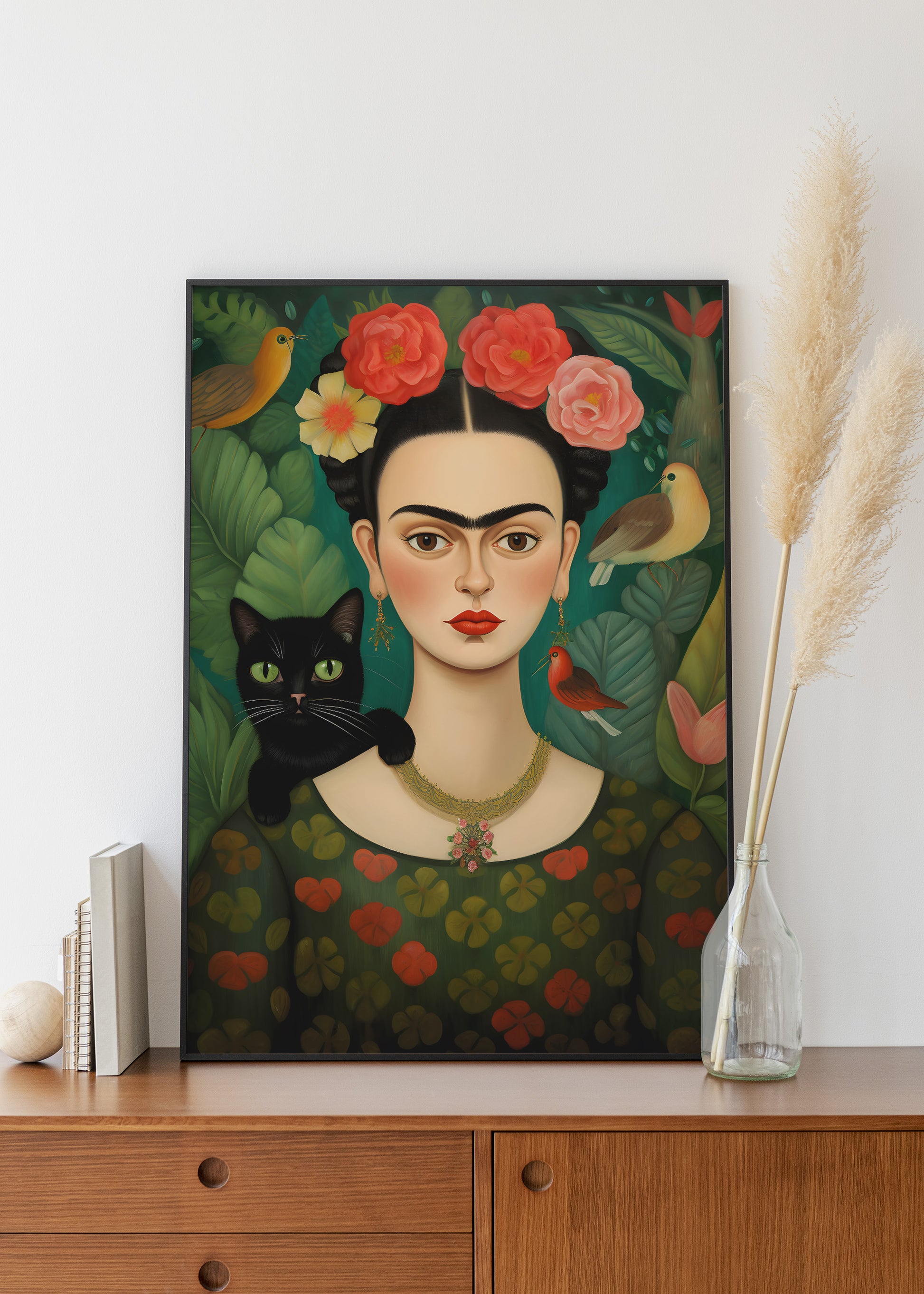 A framed art print of Frida Kahlo's iconic portrait, featuring a black cat, birds, and flowers, styled on a wooden cabinet next to a vase with pampas grass.