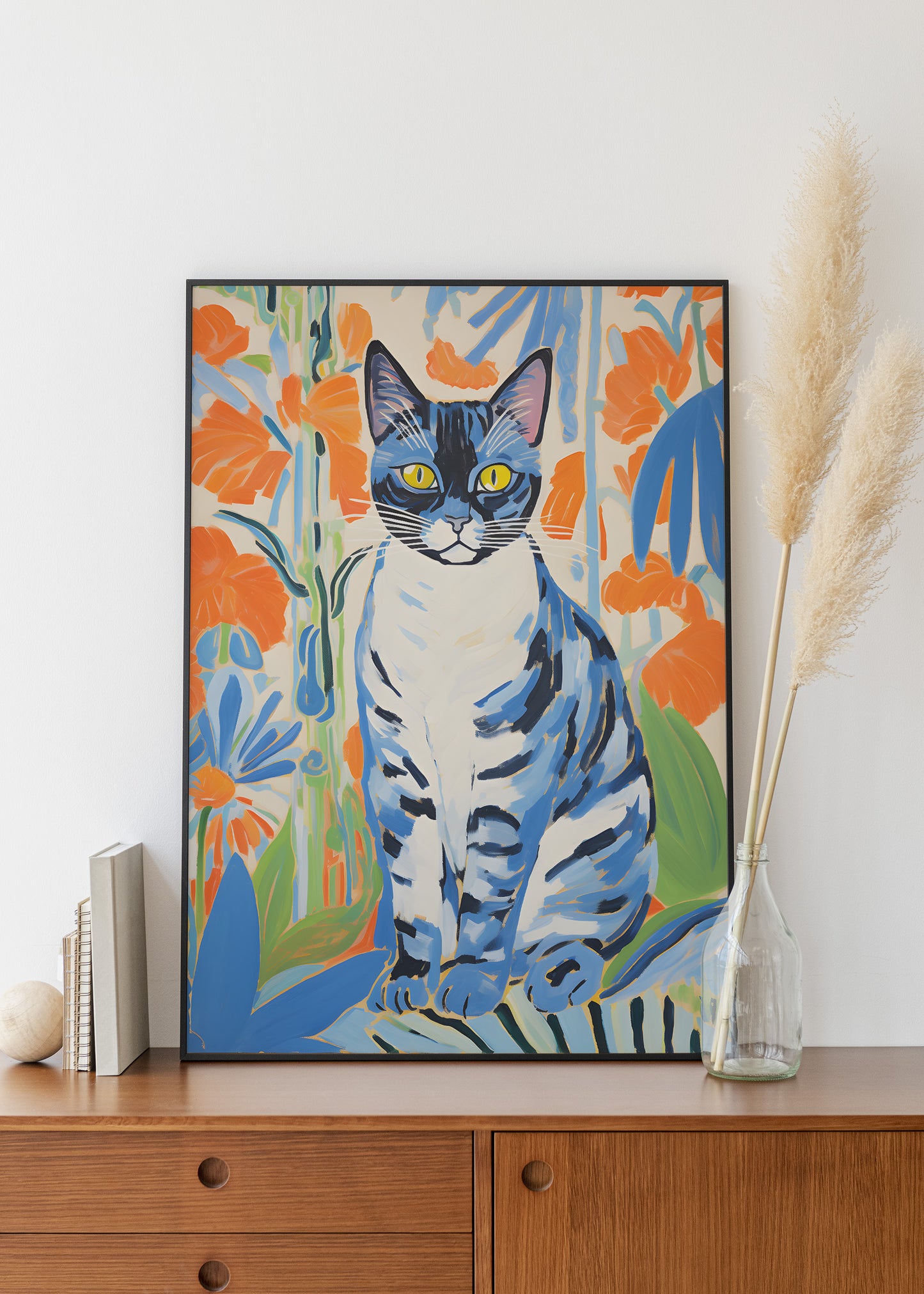 A framed art print of a blue and white tabby cat surrounded by orange flowers, displayed on a wooden cabinet next to a vase with dried pampas grass.