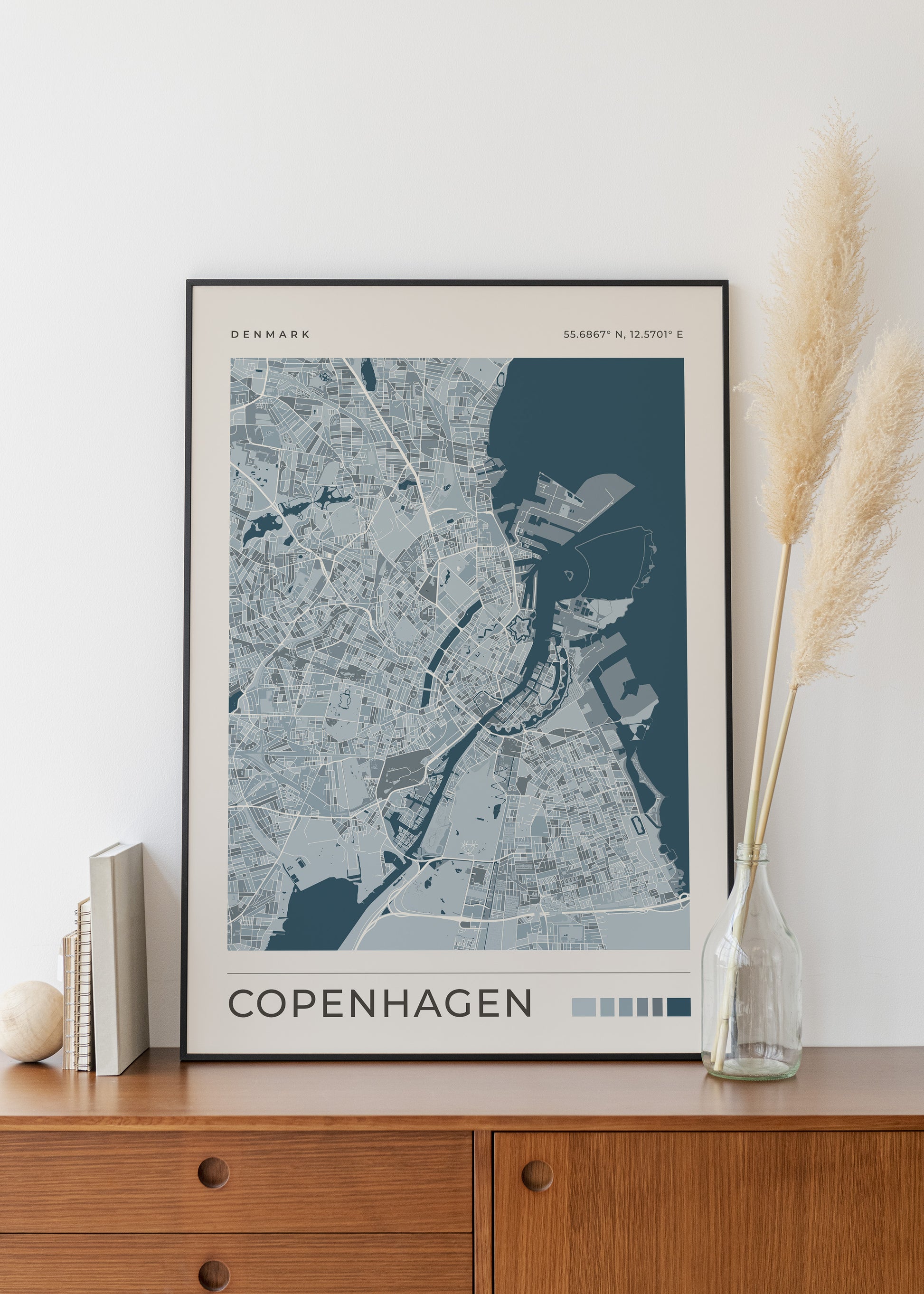 A framed Copenhagen city map print with a dark navy and light gray design, leaning on a wooden cabinet against a white wall, styled with books, a small vase, and dried pampas grass.