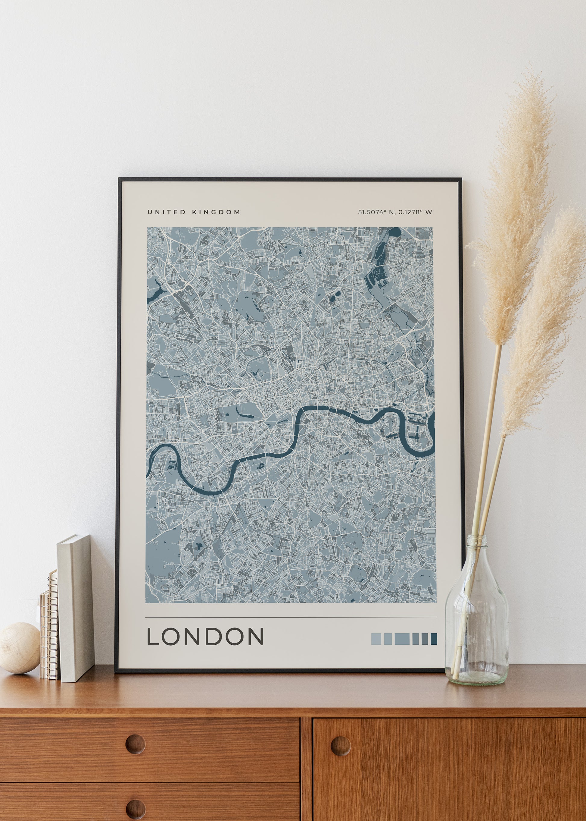 A framed London city map art print with a blue and gray street pattern, displayed on a wooden cabinet against a white wall, styled with books and a glass vase of pampas grass.