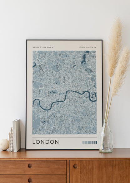 A framed London city map art print with a blue and gray street pattern, displayed on a wooden cabinet against a white wall, styled with books and a glass vase of pampas grass.