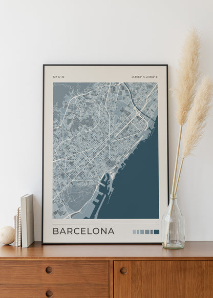 A framed Barcelona city map print leaning on a wooden cabinet against a white wall, styled with books, a small vase, and dried pampas grass.