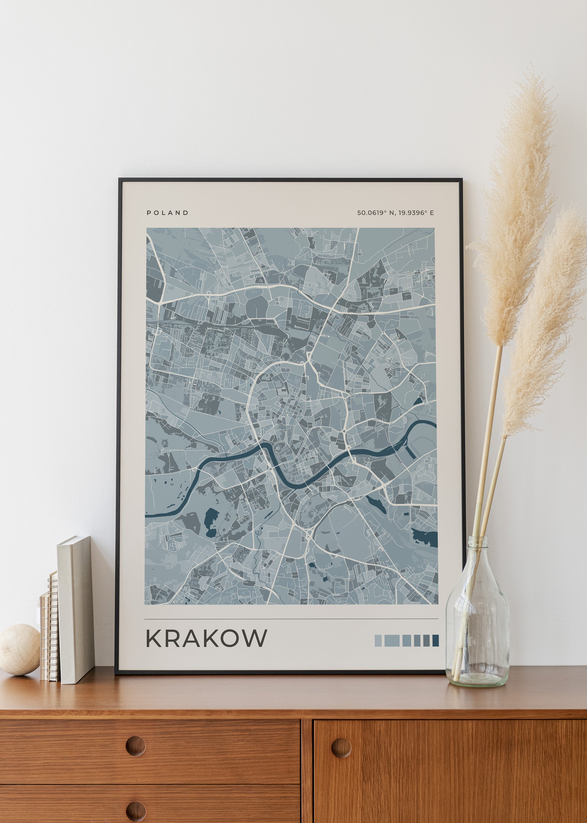 A framed minimalist city map print of Krakow, Poland, styled on a wooden sideboard next to a glass vase with pampas grass.