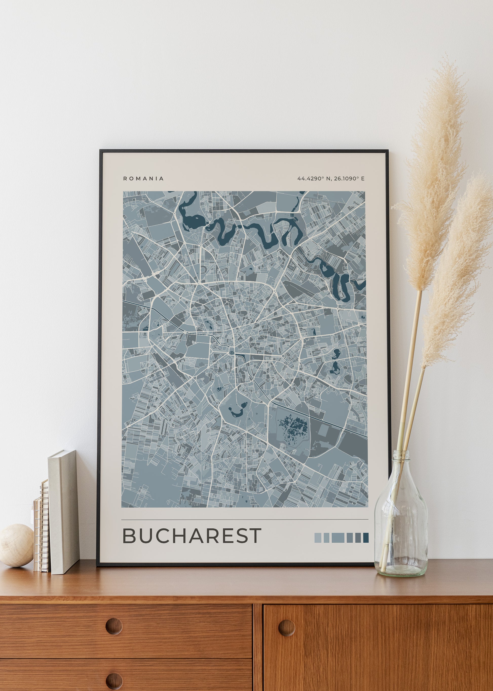 A framed city map poster of Bucharest, Romania, displayed on a wooden cabinet against a white wall, styled with books, a small vase, and pampas grass.