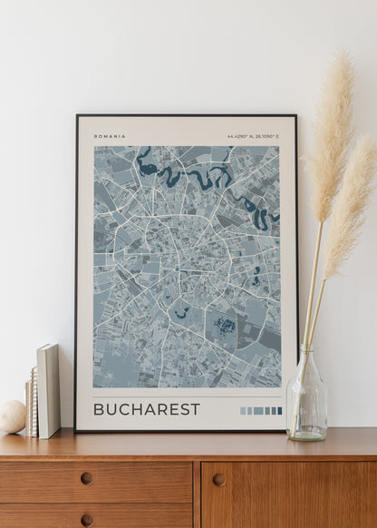 A framed city map poster of Bucharest, Romania, displayed on a wooden cabinet against a white wall, styled with books, a small vase, and pampas grass.