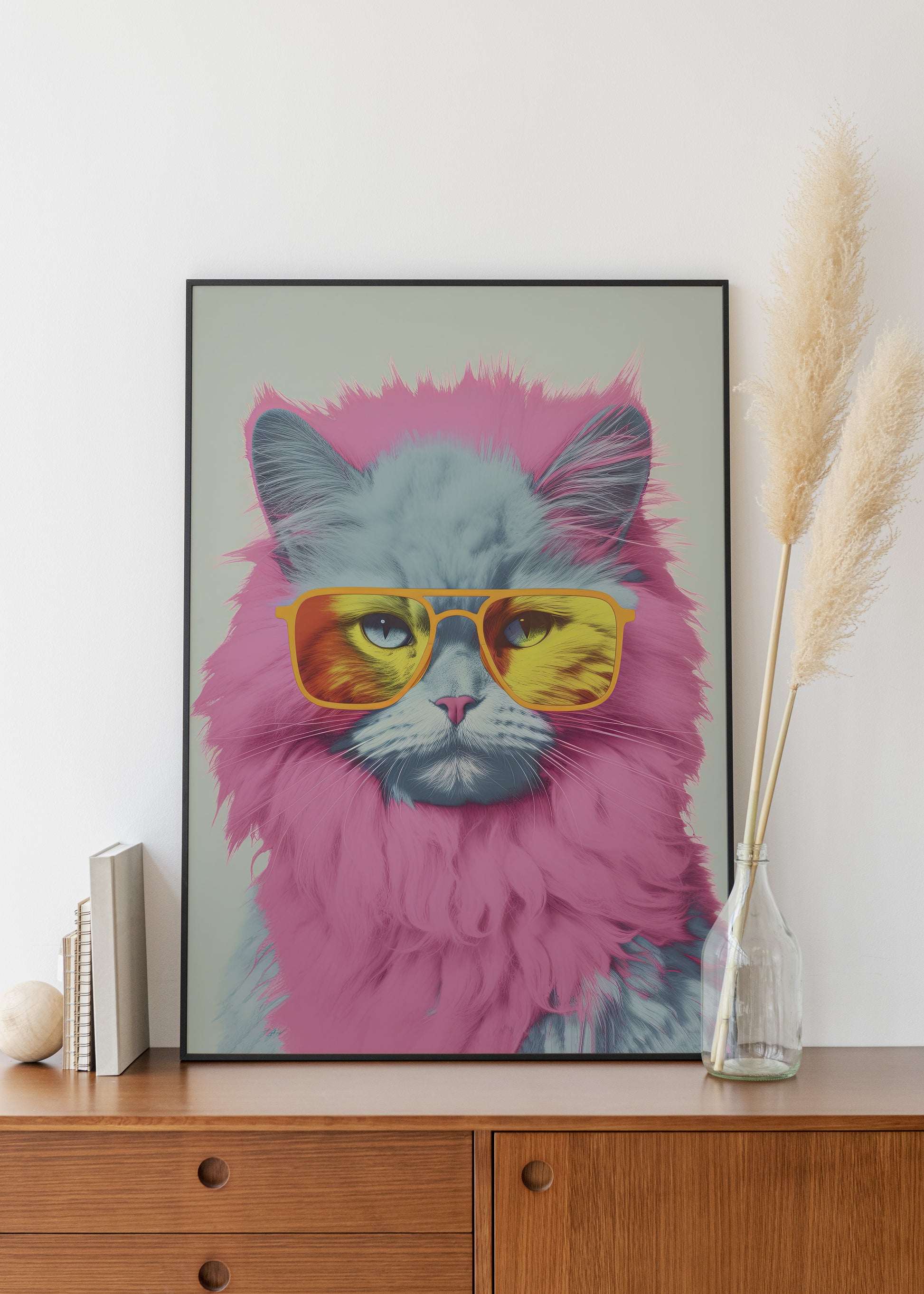 A framed art print featuring a fluffy gray cat with pink fur and yellow mirrored sunglasses, displayed on a wooden cabinet against a white wall with decorative pampas grass in a glass vase.