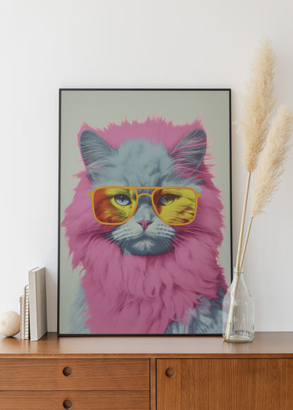A framed art print featuring a fluffy gray cat with pink fur and yellow mirrored sunglasses, displayed on a wooden cabinet against a white wall with decorative pampas grass in a glass vase.