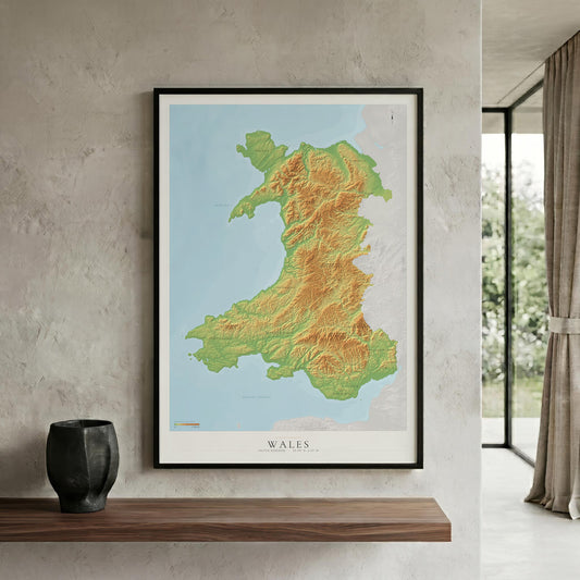 A framed topographic map poster of Wales hangs on a textured light gray wall above a wooden shelf. The print shows the UK map with green and yellow relief shading on a light blue sea background, with the word 'WALES' at the bottom.