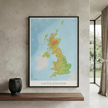 A framed topographical map poster of the United Kingdom hangs on a textured light gray wall above a wooden shelf. The poster features a light blue ocean background with green and yellow landmasses, enclosed in a thin black frame.