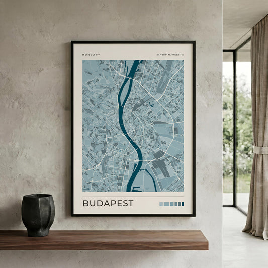 A framed Budapest city map print hangs on a textured light gray wall above a wooden shelf, with a dark vase on the shelf and a window with curtains to the right.