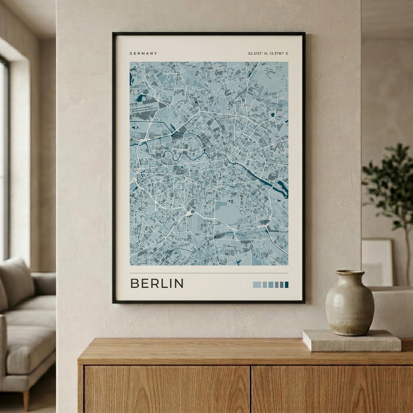 A framed map poster of Berlin, Germany, featuring a light blue and white street layout with a black frame, displayed on a beige wall above a wooden console table in a modern living room setting.