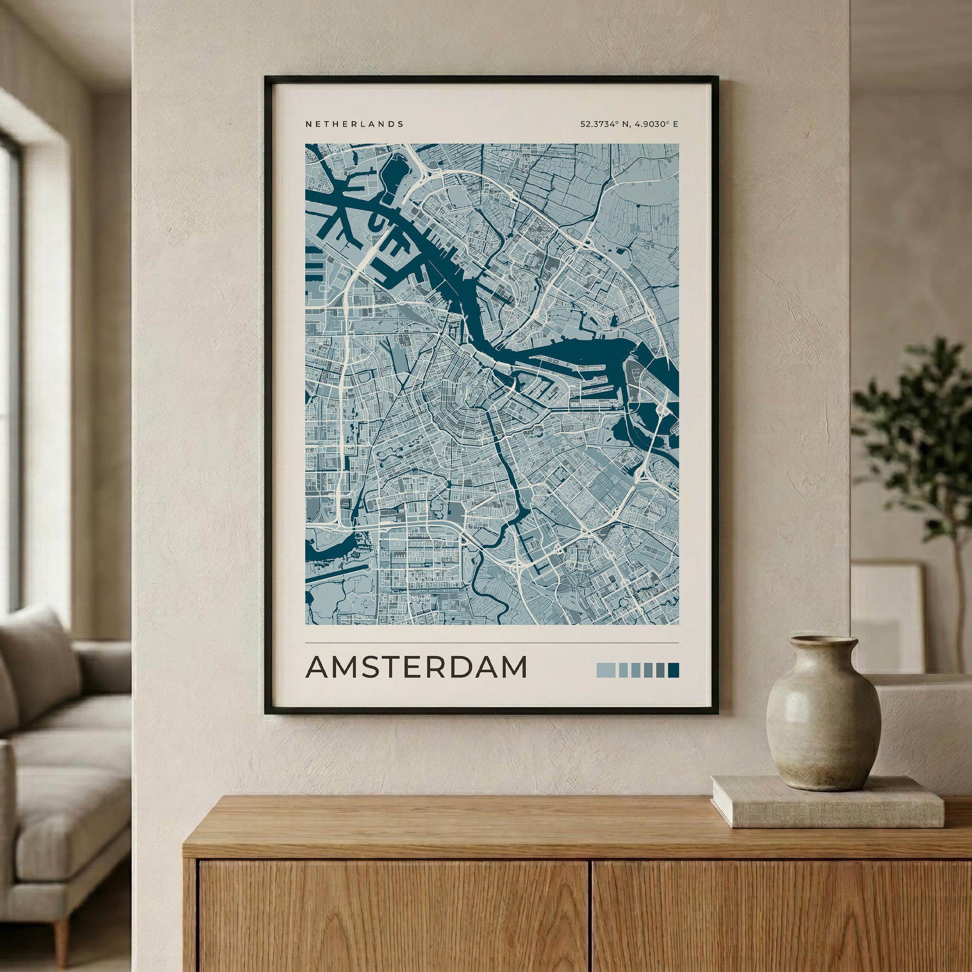 A framed Amsterdam city map print with blue and white street details, hanging on a beige wall above a wooden console table in a modern living room setting.