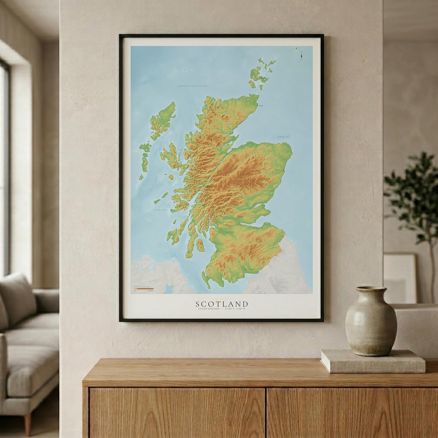 A framed Scotland topographic map print hangs on a beige wall above a wooden console table, with a vase and part of a living room visible in the background.