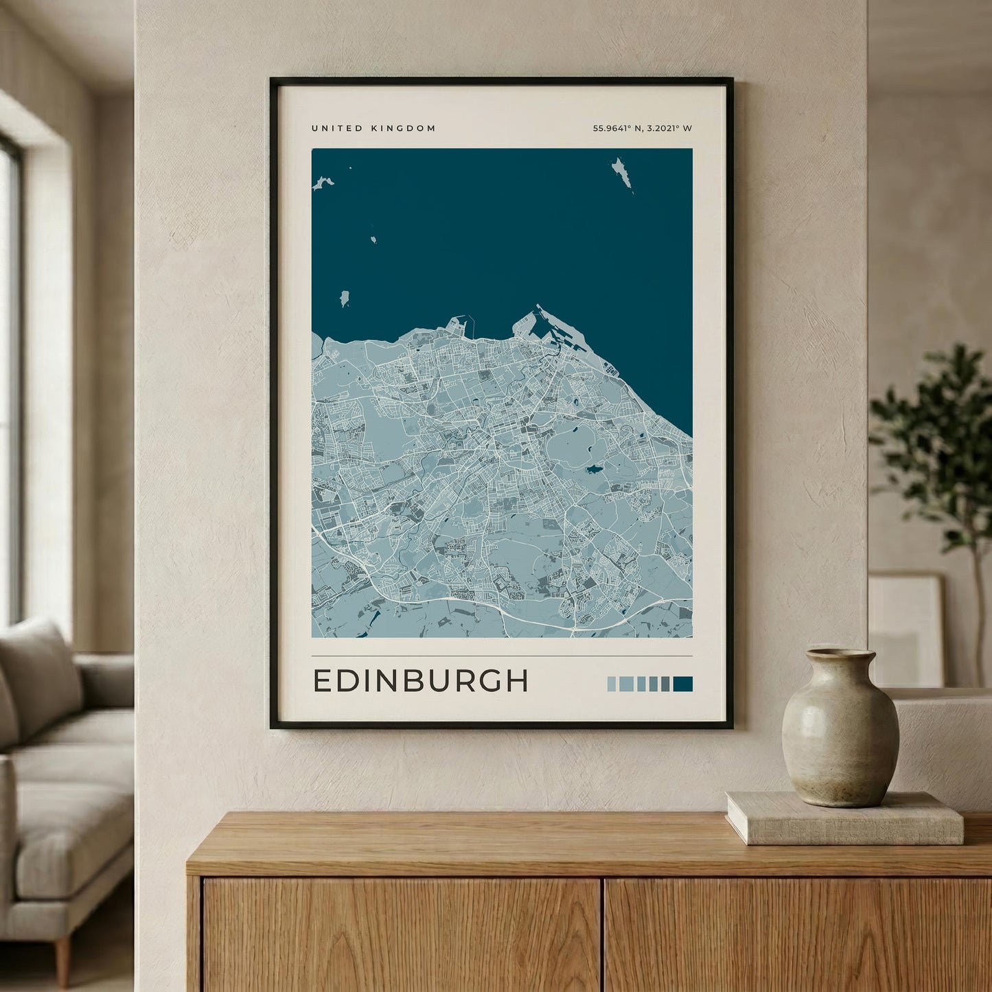 A framed Edinburgh city map print hangs on a beige wall above a wooden console table in a modern living room setting. The map features a dark teal ocean and light blue landmass with the word 'EDINBURGH' printed at the bottom.