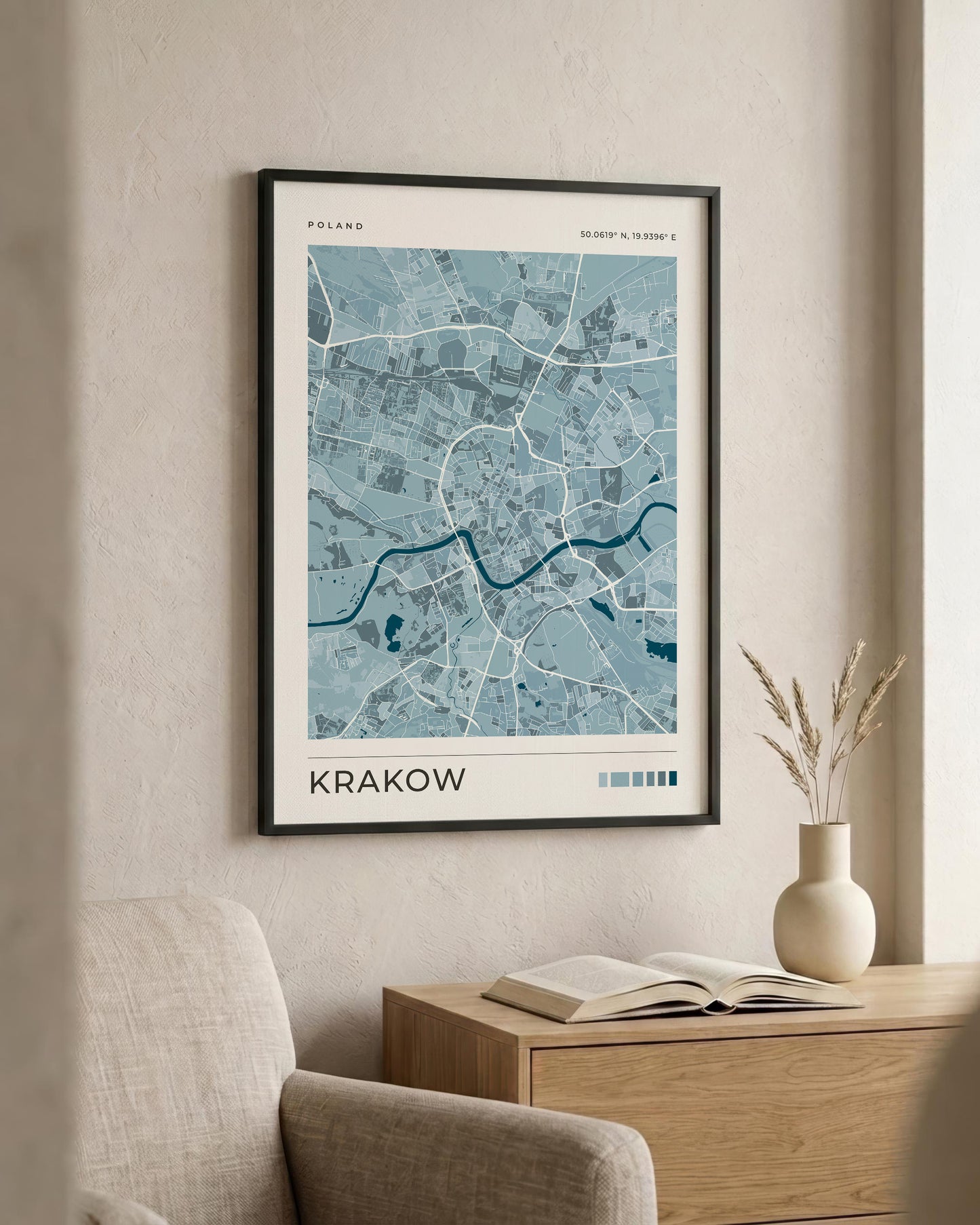 A framed city map print of Krakow, Poland, featuring a light blue and white street layout with a dark blue river, displayed on a beige wall above a wooden side table with a vase and book.