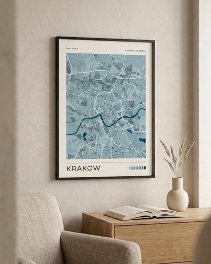 A framed city map print of Krakow, Poland, featuring a light blue and white street layout with a dark blue river, displayed on a beige wall above a wooden side table with a vase and book.