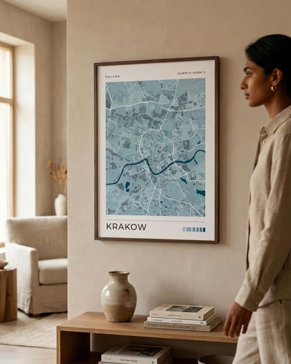 A framed blue map print of Krakow hangs on a beige wall in a modern living room setting, with a woman in a beige shirt standing to the right of the frame.