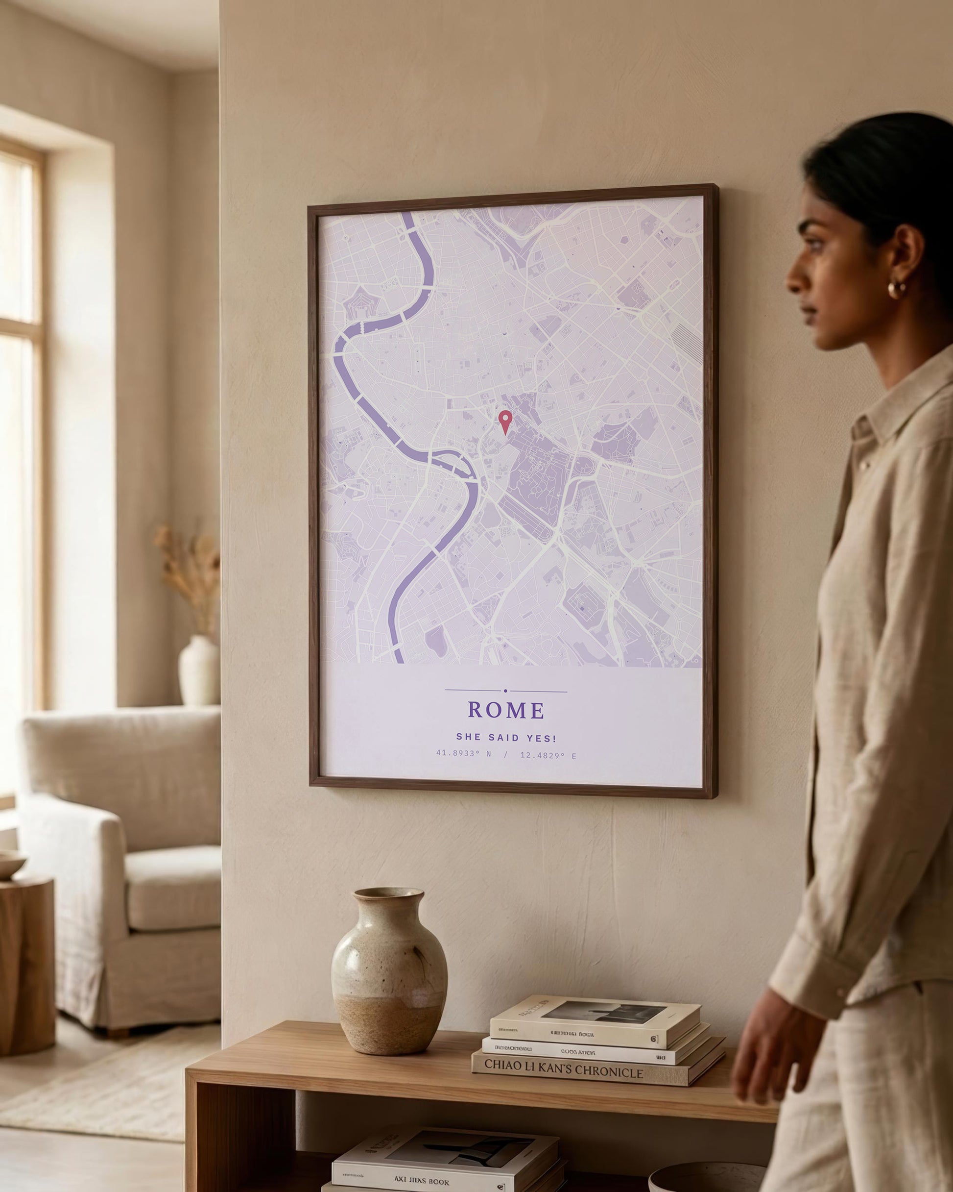 A woman in a beige outfit stands beside a framed art print of a Rome street map hanging on a beige wall in a modern, sunlit living room.