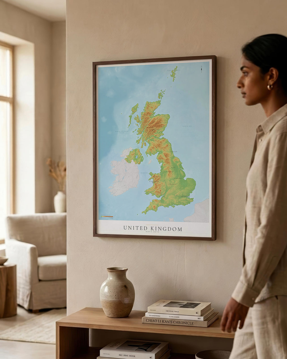 A framed topographic map poster of the United Kingdom hangs on a beige wall in a modern, sunlit room. A woman in a beige linen outfit stands to the right, looking at the print.