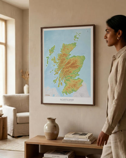 A framed Scotland topographic map print hangs on a beige wall in a styled living room, with a woman in a beige shirt standing to the right and a wooden shelf with books and a vase below.