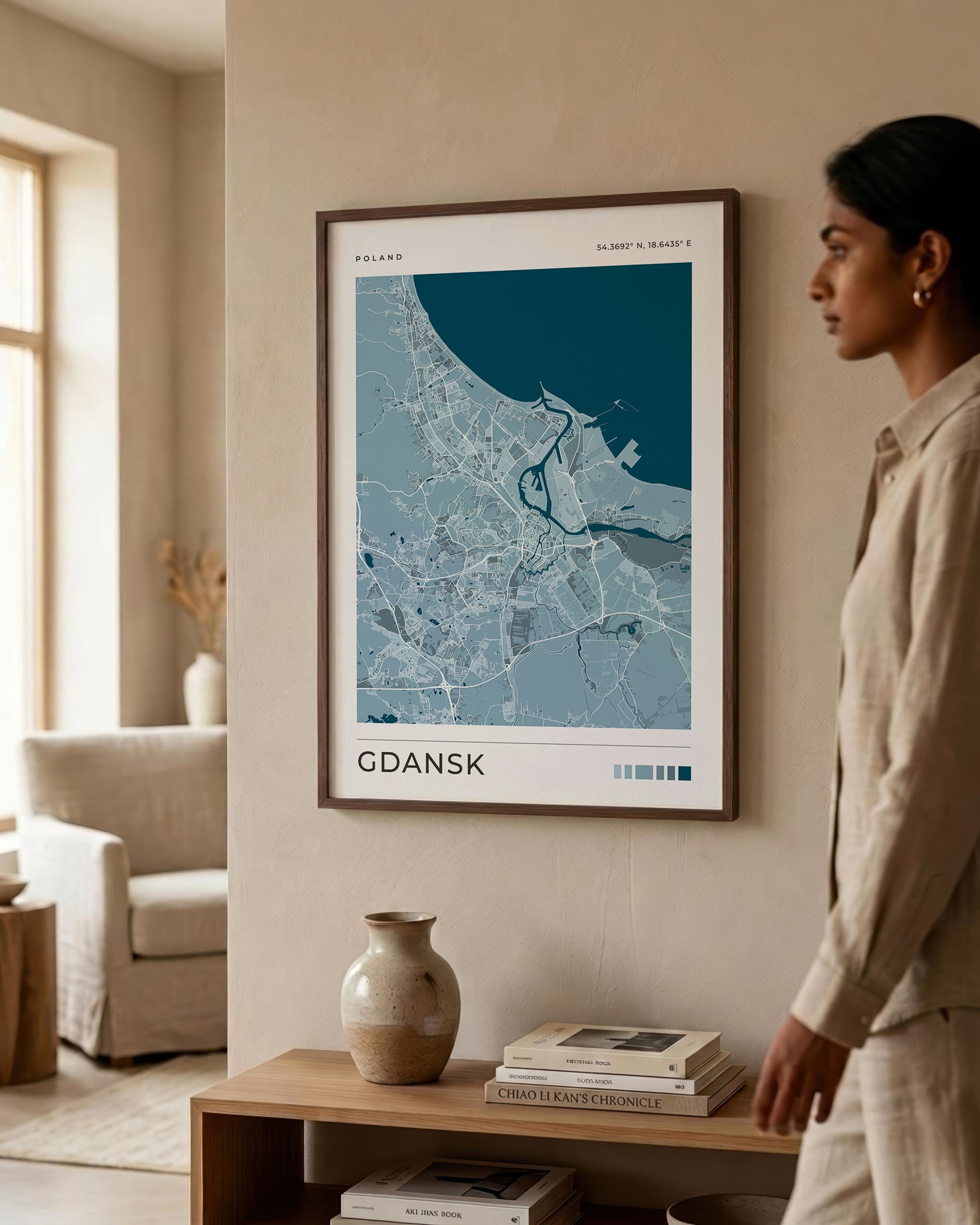 A framed art print of a stylized map of Gdansk, Poland, hanging on a beige wall in a modern living room setting. A woman in a beige linen outfit is standing to the right of the frame, looking at the print.