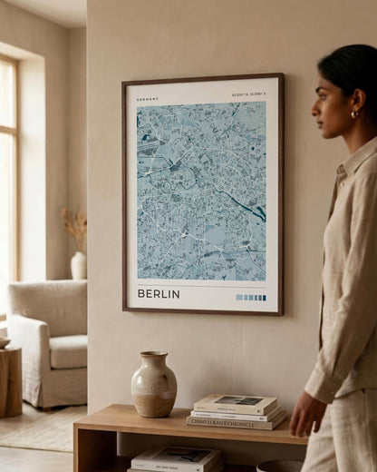 A framed poster of a light blue map of Berlin, Germany, hanging on a beige wall in a modern, sunlit living room. A woman in a beige linen outfit is standing to the right of the frame, looking at the artwork.