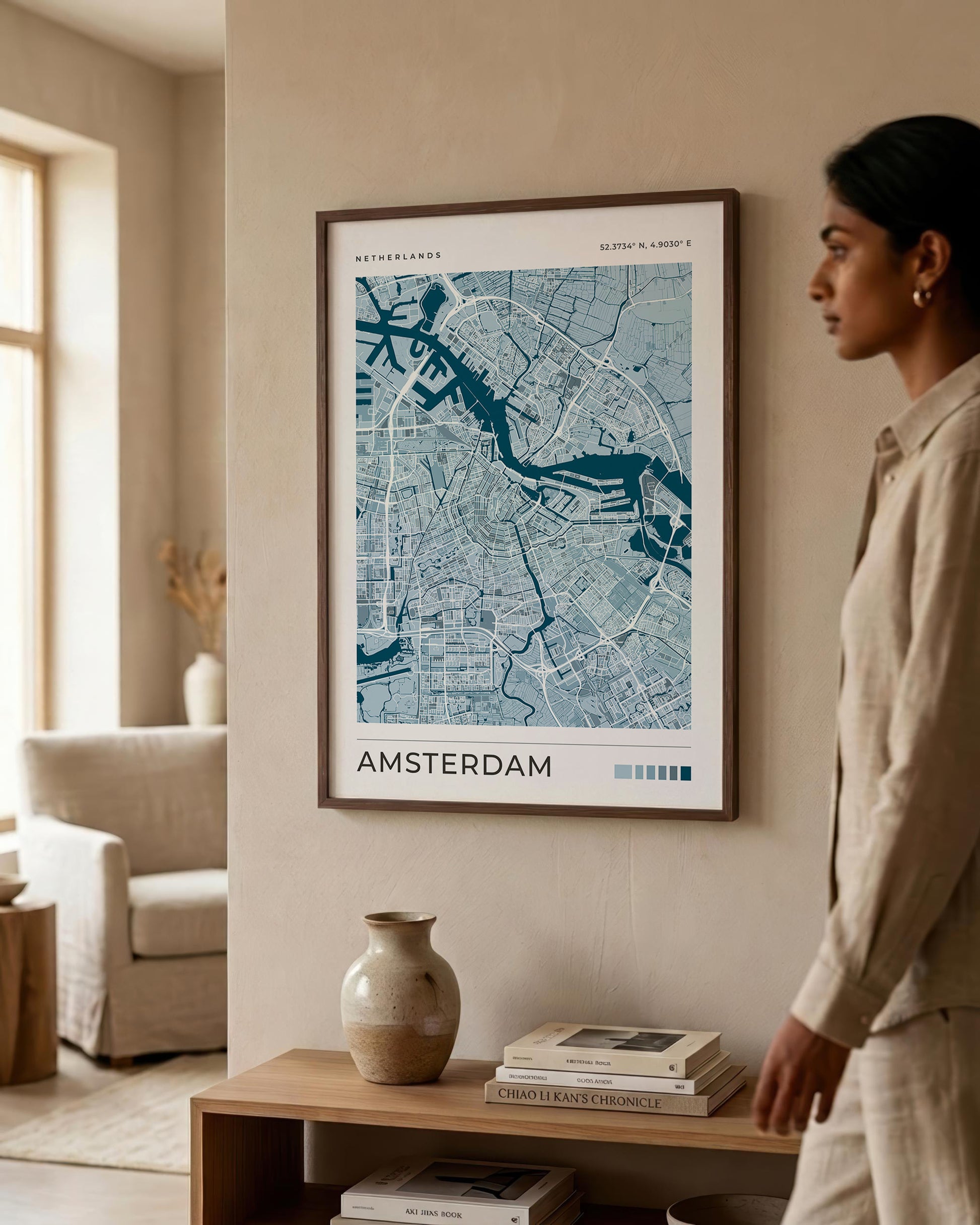 A framed blue and white Amsterdam city map print hangs on a beige wall in a modern living room, with a woman standing to the right and a wooden shelf below holding books and a vase.