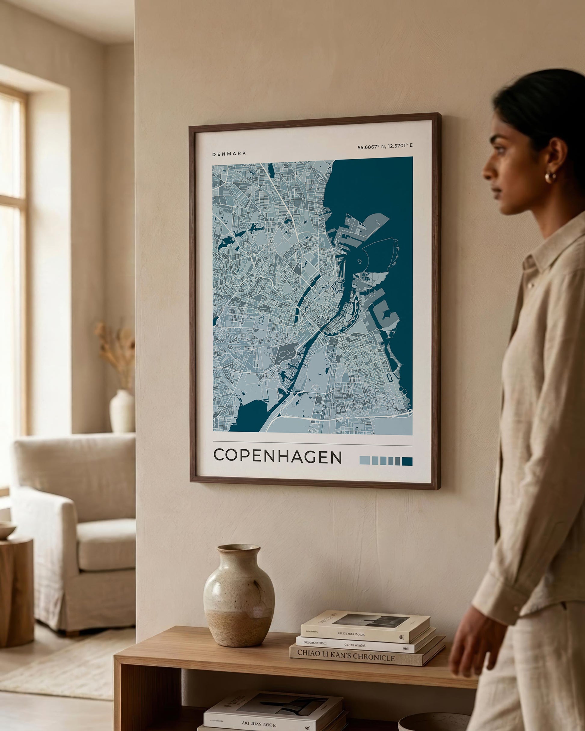 A framed Copenhagen city map print hangs on a beige wall in a modern living room, with a woman standing to the right and a wooden console table below holding books and a vase.