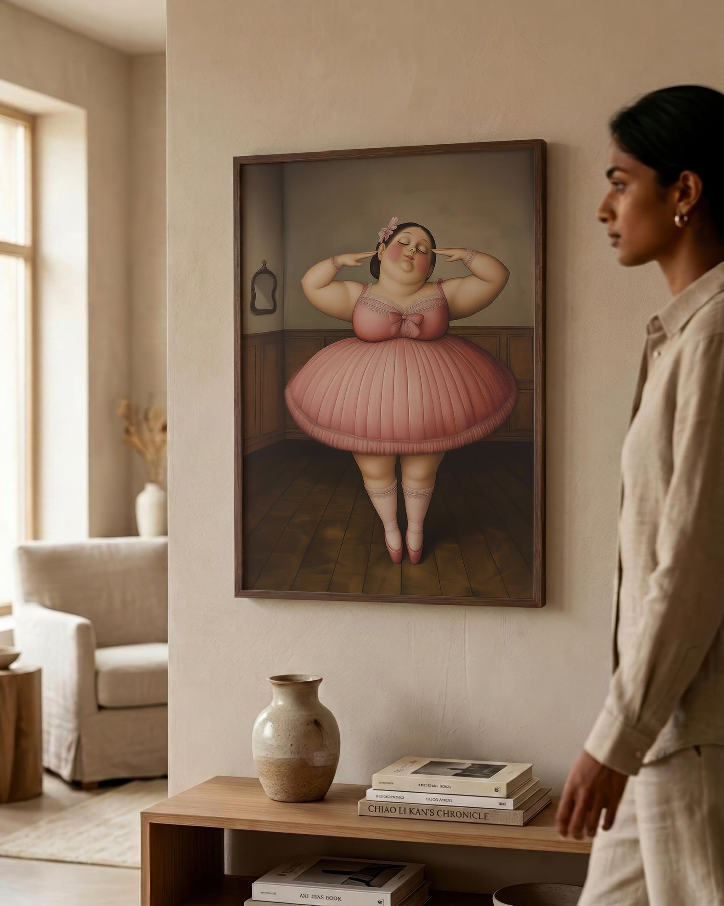 A woman in a beige linen outfit stands in a modern, sunlit room looking at a framed art print on the wall. The print depicts a stylized, painterly portrait of a woman in a pink tutu holding a pink flower to her ear.