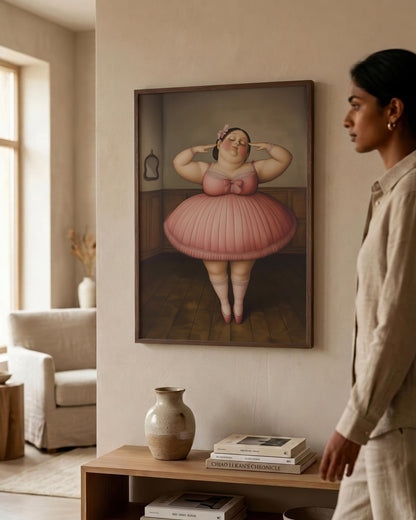 A woman in a beige linen outfit stands in a modern, sunlit room looking at a framed art print on the wall. The print depicts a stylized, painterly portrait of a woman in a pink tutu holding a pink flower to her ear.