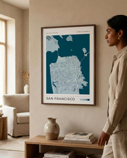 A framed San Francisco city map poster hangs on a beige wall in a styled living room. A woman in a beige outfit stands to the right, looking at the artwork.