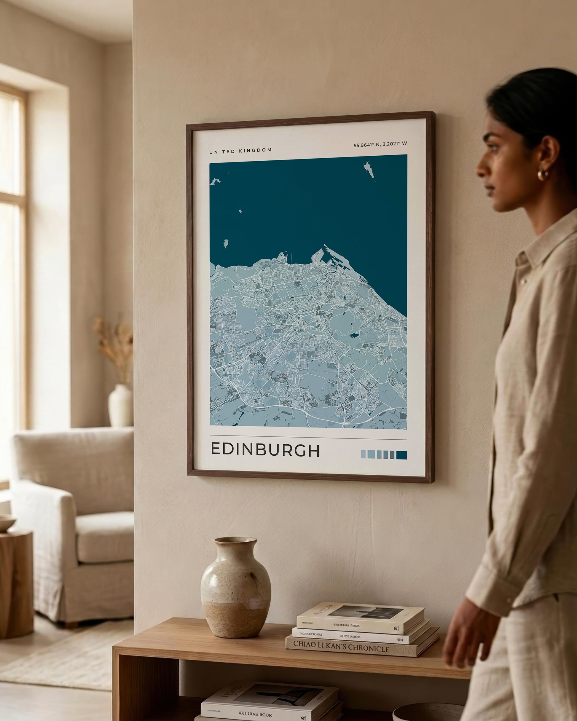A framed Edinburgh city map print hangs on a beige wall in a modern living room, with a woman standing to the right and a wooden console table below holding books and a vase.