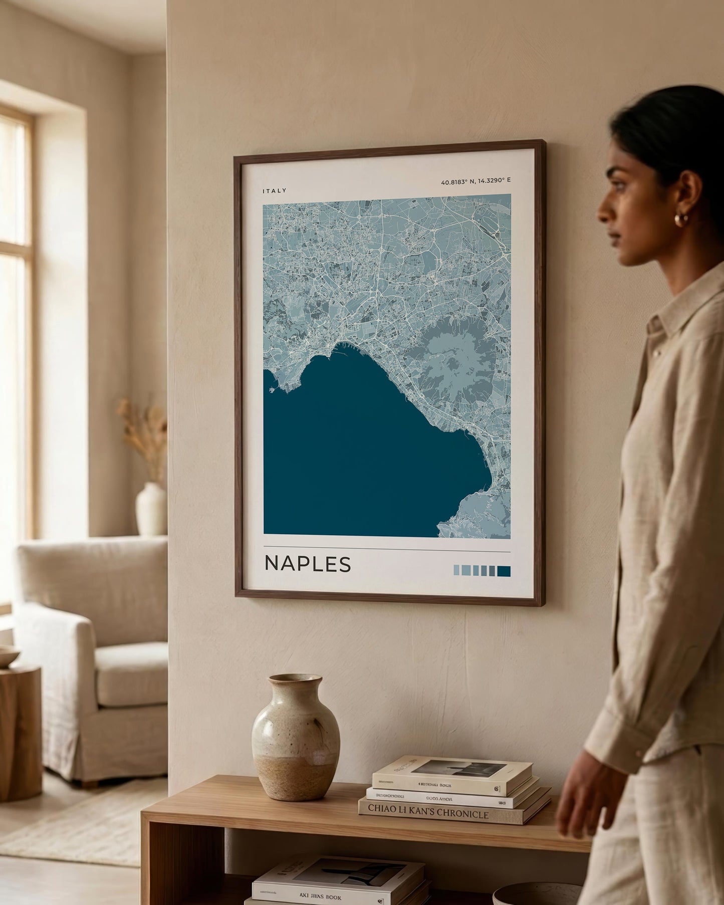 A framed art print of a Naples map hangs on a beige wall in a modern, sunlit room. A woman in a beige linen outfit stands to the right, looking at the print.