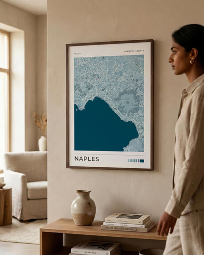 A framed art print of a Naples map hangs on a beige wall in a modern, sunlit room. A woman in a beige linen outfit stands to the right, looking at the print.
