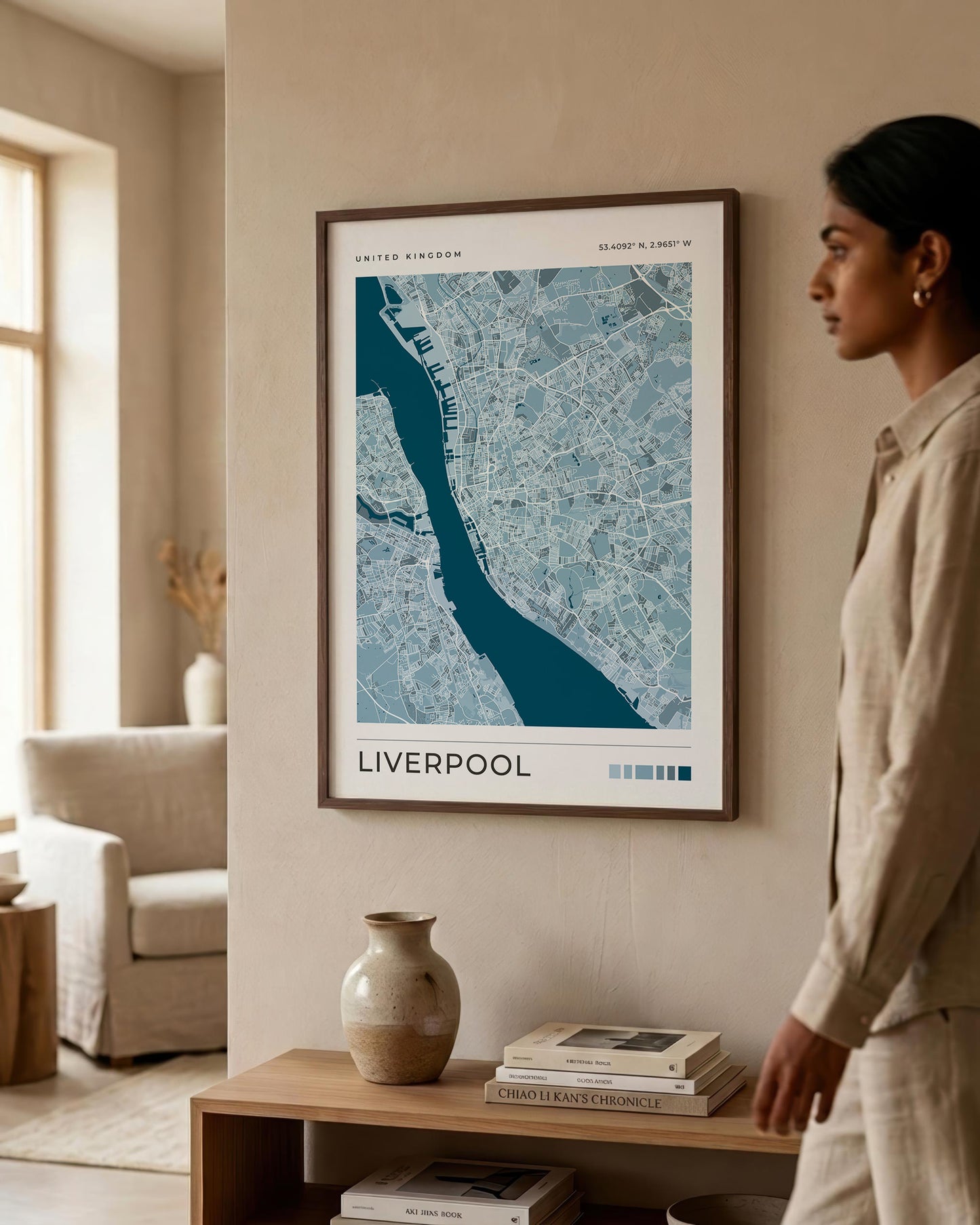A framed map print of Liverpool, United Kingdom, displayed on a beige wall in a modern living room setting. A woman in a beige linen outfit is standing to the right of the frame, looking at the artwork.