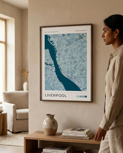 A framed map print of Liverpool, United Kingdom, displayed on a beige wall in a modern living room setting. A woman in a beige linen outfit is standing to the right of the frame, looking at the artwork.