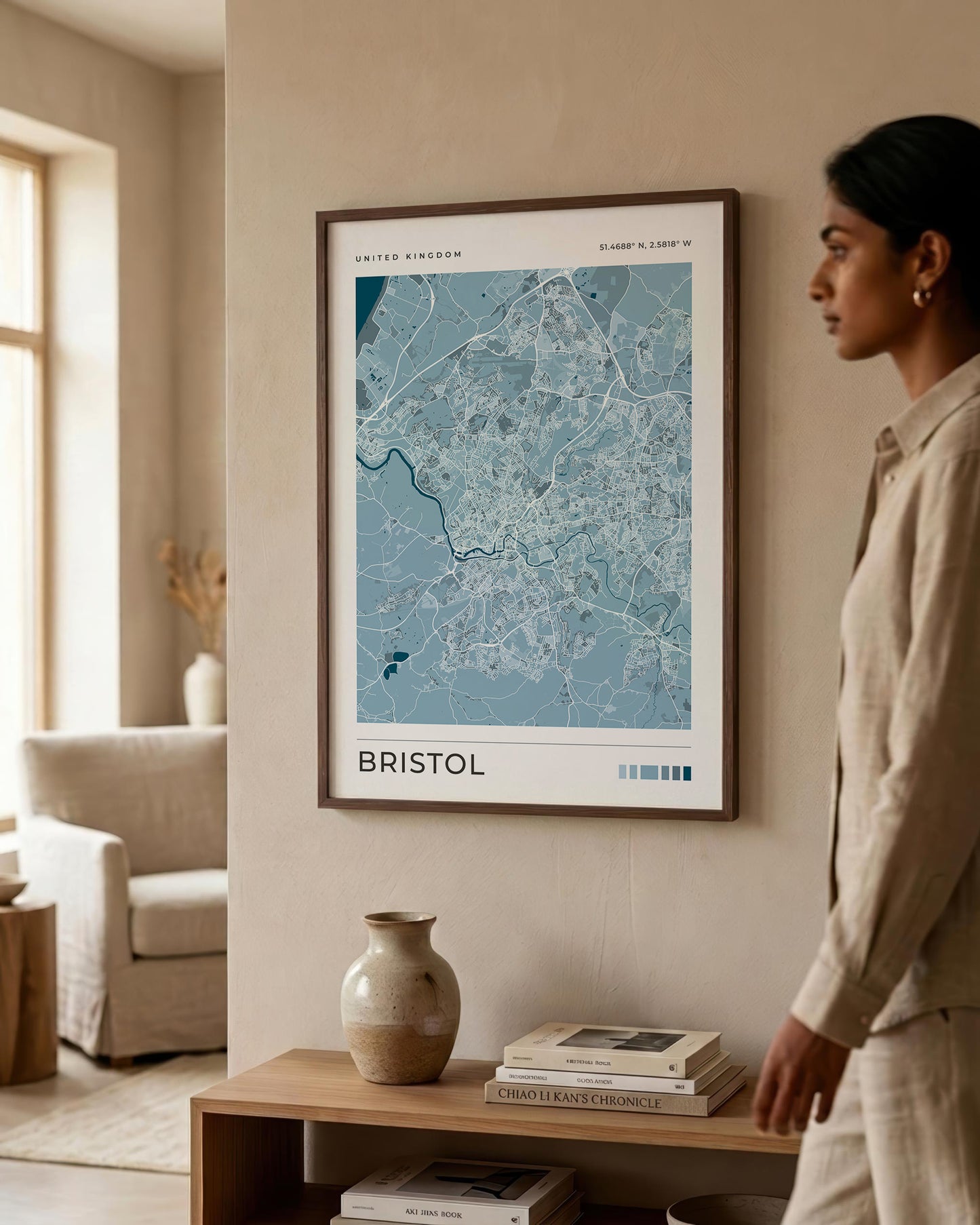 A framed city map print of Bristol, United Kingdom, hanging on a beige wall in a modern, sunlit living room. A woman in a beige linen outfit stands to the right, looking at the artwork.