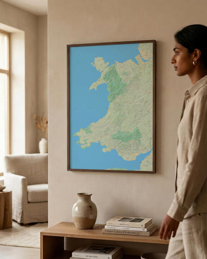 A framed topographic map of Wales hanging on a beige wall in a modern living room, with a woman in a beige linen outfit standing to the right and a wooden console table below with books and a ceramic vase.