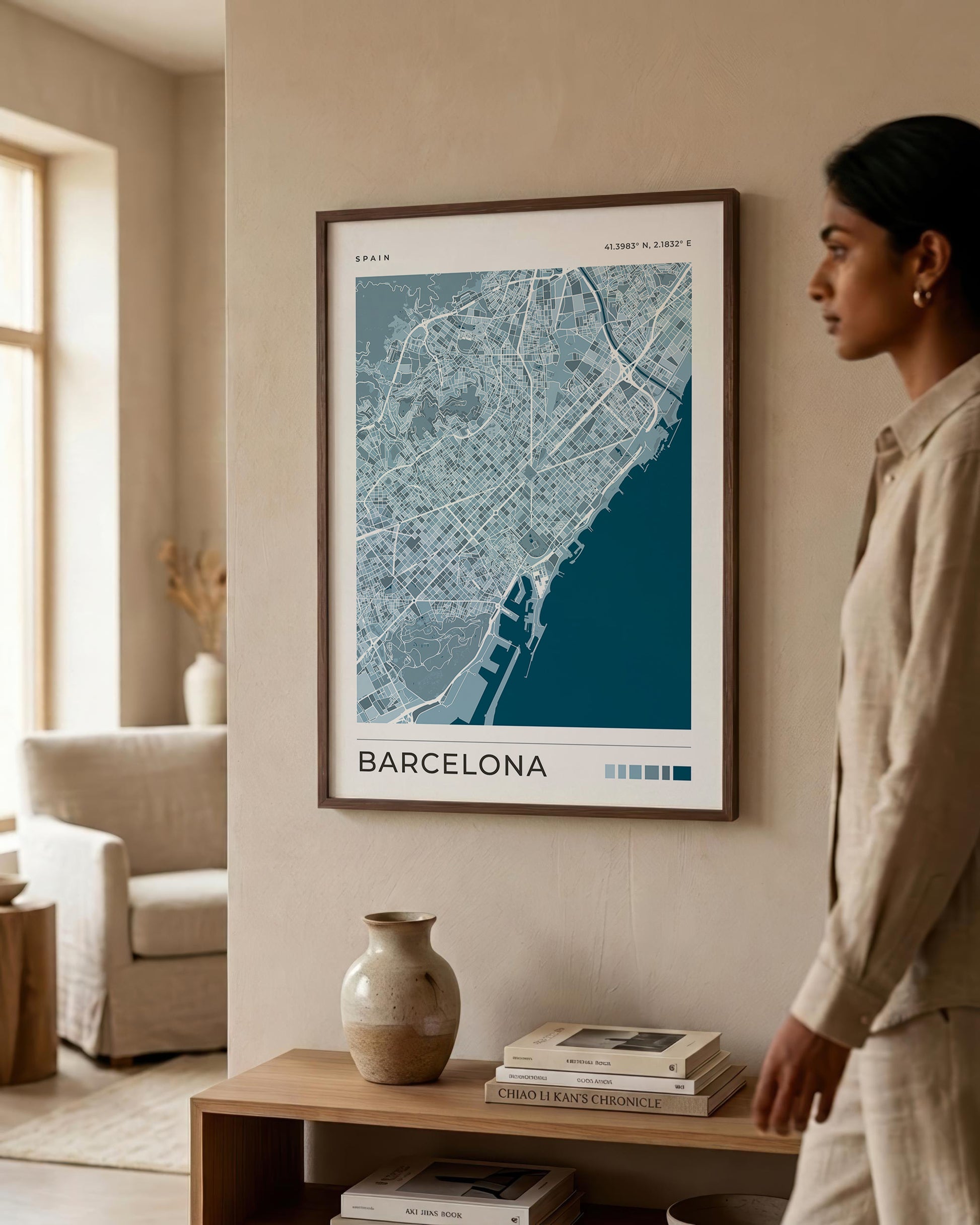A framed Barcelona city map print hangs on a beige wall in a modern living room, with a woman standing beside it and a wooden console table below holding books and a vase.